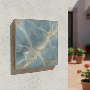 May include: A square, blue-grey decorative tile with a marble-like pattern of gold and white veining. The tile is mounted on a white wall, casting a shadow. The overall aesthetic is modern and elegant.
