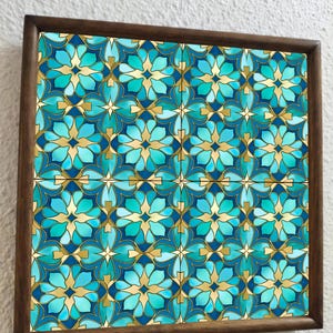 May include: Square wooden framed artwork featuring a repeating floral pattern in shades of teal, turquoise, and gold. The design is symmetrical and geometric, with a dark brown wooden frame. The artwork is mounted on a white wall.