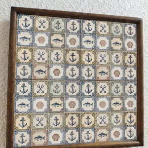 May include: A framed tile art piece featuring nautical designs. The tiles are decorated with anchors, fish, seashells, and crossed oars in shades of blue, green, and beige. The frame is made of dark brown wood.