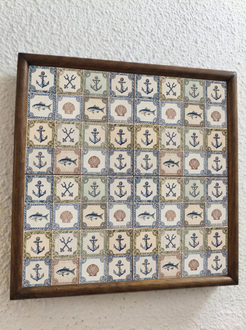 May include: A framed tile art piece featuring nautical designs. The tiles are decorated with anchors, fish, seashells, and crossed oars in shades of blue, green, and beige. The frame is made of dark brown wood.