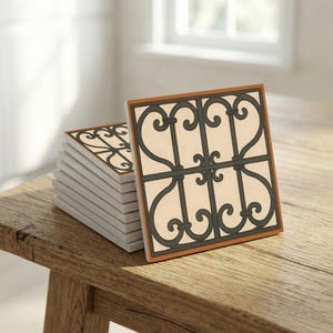 Puede incluir: Una pila de posavasos cuadrados de cerámica con un diseño decorativo de hierro forjado negro sobre un fondo crema. Los posavasos tienen un borde marrón y se exhiben sobre una superficie de madera.