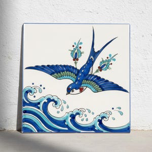 Puede incluir: Un azulejo de cerámica cuadrado con una golondrina azul pintada a mano en vuelo sobre olas oceánicas estilizadas en azul y turquesa. El ave presenta detalles en blanco, azul y rojo, con acentos florales decorativos tras las alas sobre fondo blanco.
