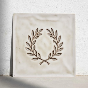 May include: Square, off-white ceramic tile featuring a detailed, brown laurel wreath design. The wreath is centered on the tile, with the leaves forming a circular pattern. The tile has a slightly textured surface.