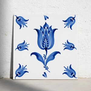 May include: A square ceramic tile with a white background featuring a large central blue tulip illustration surrounded by six smaller blue tulip motifs arranged in a symmetrical pattern. The design uses varying shades of blue paint.