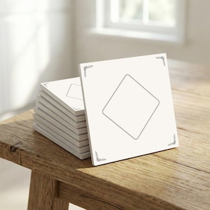 Puede incluir: Una pila de posavasos cuadrados blancos con un diseño de rombo gris en el centro y detalles decorativos en las esquinas. Los posavasos se exhiben sobre una mesa de madera rústica, iluminada por luz natural.