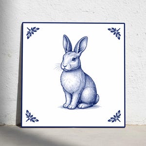 Puede incluir: Azulejo de cerámica cuadrado con una ilustración azul y blanca de un conejo sentado. La obra de arte tiene un aspecto dibujado a mano. Motivos decorativos de hojas en cada esquina. El azulejo está sobre un fondo blanco.