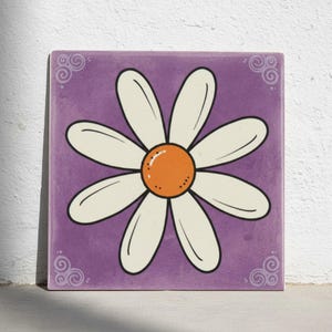 May include: Square ceramic tile with a daisy design. The daisy has white petals with black outlines and an orange centre. The background is purple, and the corners have decorative swirl patterns. The tile is likely for home decor.
