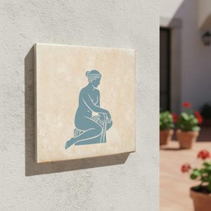 Puede incluir: Azulejo cuadrado beige con una silueta azul de una figura sentada. La figura está cubierta con una tela y está sentada en un taburete. El azulejo está montado en una pared de color claro, lo que sugiere un entorno al aire libre.