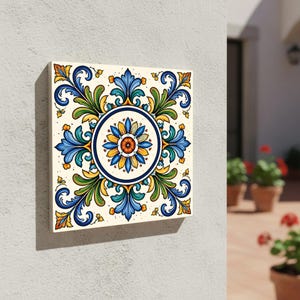 Puede incluir: Azulejo cerámico cuadrado con diseño floral. El azulejo presenta una flor central con pétalos naranjas, amarillos y azules, rodeada de hojas azules y verdes. El fondo es blanquecino, y el azulejo está montado en una pared blanca.