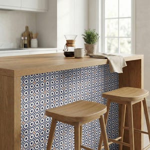 Op de afbeelding: Houten keukeneiland met een patroon van tegels in marineblauw, wit en oranje. Twee houten krukken staan ervoor. Een koffiezetapparaat, een mok en een plant staan op het eiland. Het patroon herhaalt zich.