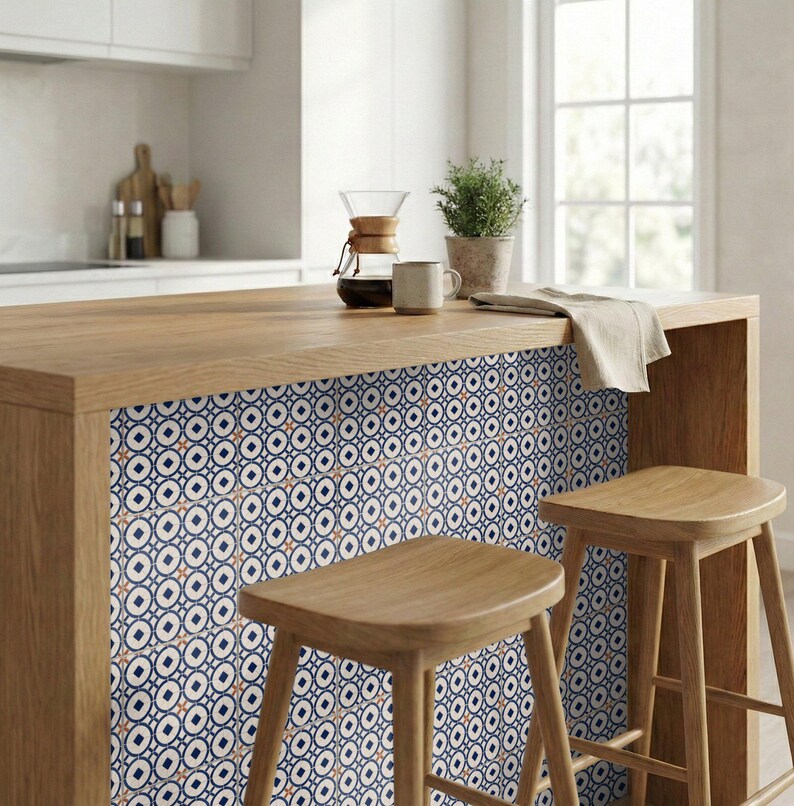 Op de afbeelding: Houten keukeneiland met een patroon van tegels in marineblauw, wit en oranje. Twee houten krukken staan ervoor. Een koffiezetapparaat, een mok en een plant staan op het eiland. Het patroon herhaalt zich.