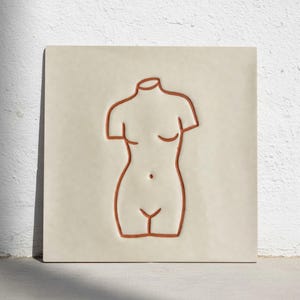 Puede incluir: Una baldosa de cerámica cuadrada de color crema con un contorno minimalista de un torso en un tono marrón rojizo cálido. La obra de arte representa la parte superior del cuerpo con líneas simplificadas, mostrando las curvas y los detalles de la forma.