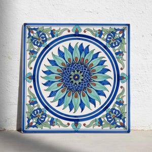 May include: A square ceramic tile featuring a central circular medallion with a blue and turquoise floral motif. The design includes intricate petal patterns, a dark blue grid center, and decorative corner elements in shades of blue and green.