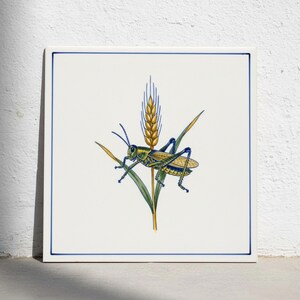 Puede incluir: Un azulejo de cerámica cuadrado con fondo blanco y un borde azul fino. El centro presenta una ilustración detallada de un saltamontes verde y amarillo posado sobre un tallo de trigo dorado con hojas verdes largas, en un estilo clásico.