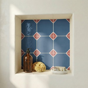 Op de afbeelding: Een badkamisnis met blauw en rood betegelde patronen. Een bruine glazen fles met een pompdosering, een natuurlijke spons en een klein wit schaaltje met zeep staan op de plank. De nis wordt omlijst door witte muren.