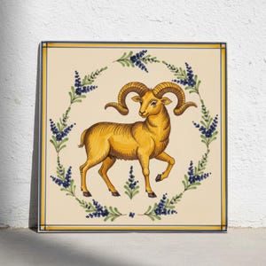 May include: A square ceramic tile featuring a central illustration of a golden-brown ram with curved horns. The ram is encircled by a decorative wreath of green stems and small blue flowers. The tile has a cream background and a blue and yellow border.