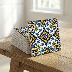 Può includere: Una pila di sottobicchieri quadrati in ceramica con un motivo floreale ripetuto in blu navy, giallo e bianco. I sottobicchieri sono esposti su un tavolo di legno rustico. Misurano circa 10 cm x 10 cm.
