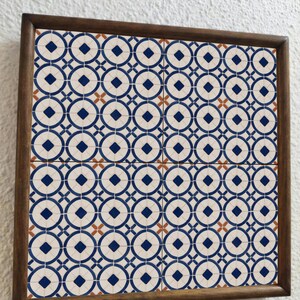 Op de afbeelding: Vierkant ingelijste tegelkunst met een herhalend patroon van blauwe cirkels, ruiten en kruisen op een cr&egrave;mekleurige achtergrond. De tegels zijn omlijst in een donkerbruine houten lijst. Het ontwerp doet denken aan vintage tegelwerk.