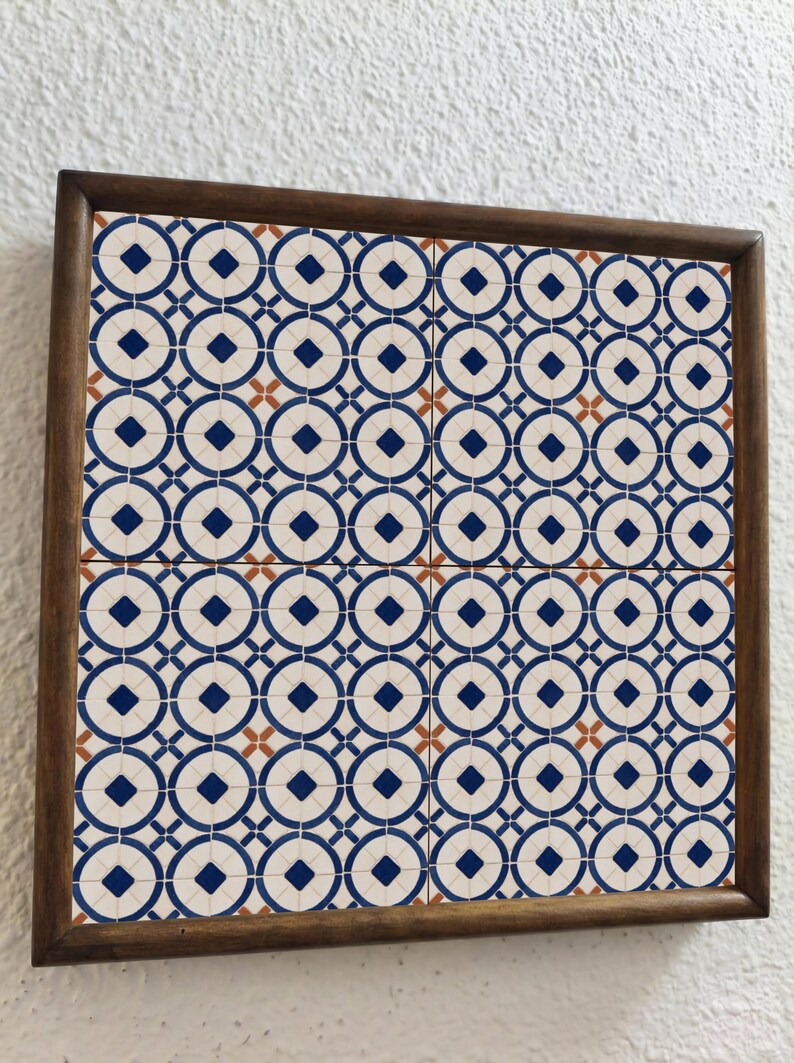 Op de afbeelding: Vierkant ingelijste tegelkunst met een herhalend patroon van blauwe cirkels, ruiten en kruisen op een cr&egrave;mekleurige achtergrond. De tegels zijn omlijst in een donkerbruine houten lijst. Het ontwerp doet denken aan vintage tegelwerk.