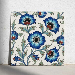 May include: A square ceramic tile featuring a repeating floral pattern on an off-white background. The design displays large blue flowers with dark centers, red accents, and light green leaves connected by thin, flowing stems and vines.