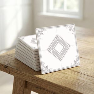 Könnte beinhalten: Ein Stapel weißer quadratischer Fliesen mit einem grauen geometrischen Design. Jede Fliese hat eine zentrale Raute und dekorative Eckelemente. Die Fliesen sind auf einer rustikalen Holzoberfläche gestapelt, mit einem Fenster im Hintergrund.