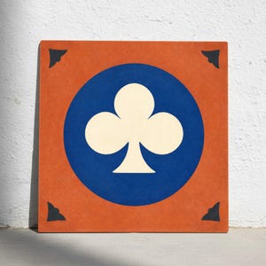 Op de afbeelding: Vierkante tegel met een klaverblad-symbool van een speelkaart. De tegel heeft een oranje achtergrond met een blauwe cirkel in het midden. Een wit klaverbladsymbool staat in het midden van de blauwe cirkel. Zwarte hoekaccenten maken het ontwerp af.