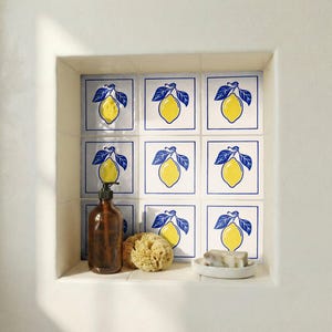 May include: A bathroom niche features square tiles with a yellow lemon and blue leaf design. A brown glass bottle with a pump, a natural sponge, and a small white dish with soap are on the shelf. The tiles create a cohesive, decorative theme.