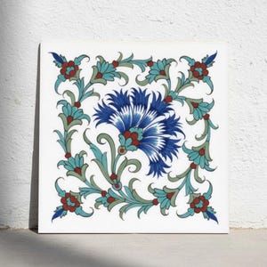 Könnte beinhalten: Eine quadratische Keramikfliese mit einer zentralen blauen Nelkenblüte, umgeben von einer symmetrischen Bordüre aus kleineren türkisfarbenen und roten Blumenmotiven. Das Design ist auf weißem Grund mit grünen Stielen und Blättern.