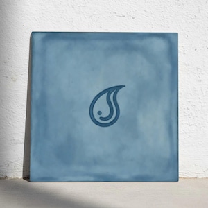 Peut inclure: Carreau bleu carré avec un motif de goutte d'eau au centre. La goutte d'eau est d'une nuance de bleu plus foncée et comporte un petit point à l'intérieur. Le carreau a un aspect texturé et est posé contre un mur blanc.