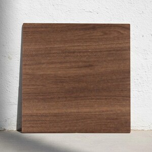 Puede incluir: Una tabla de cortar cuadrada de madera, de color marrón oscuro y con vetas visibles. La tabla se encuentra contra una pared blanca, resaltando su textura natural. Este artículo de cocina es ideal para la preparación de alimentos.