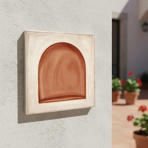 May include: A square, ceramic wall art piece featuring a terracotta-colored arch design on a cream-colored background. The artwork is framed with a thin, silver-colored metal border and mounted on a textured, light gray wall.