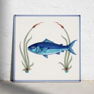 Op de afbeelding: Een vierkante keramische tegel met een centrale illustratie van een blauwe vis, geflankeerd door twee groene waterplanten met rode uiteinden. Het ontwerp is omlijst door een dunne blauwe rand op een witte achtergrond voor een strakke compositie.