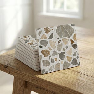 Könnte beinhalten: Ein Stapel quadratischer Fliesen mit Terrazzo-Muster. Die Fliesen zeigen ein Muster aus beigen, grauen und braunen Steinen auf weißem Grund. Die Fliesen sind auf einer Holzoberfläche gestapelt.