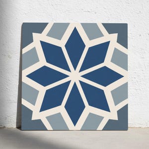 May include: Square tile with a geometric starburst design. The pattern features a central eight-pointed star in shades of blue, surrounded by cream and grey shapes. The tile is displayed against a white wall.