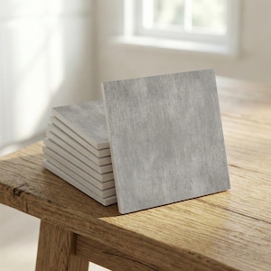 Puede incluir: Una pila de baldosas cuadradas grises con una superficie texturizada. Las baldosas están dispuestas sobre una mesa de madera rústica, con una baldosa apoyada contra la pila. Las baldosas tienen un aspecto similar al hormigón.