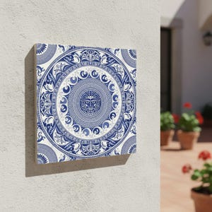 May include: Square wall art with a detailed blue and white circular design. The artwork features a central face motif surrounded by intricate patterns and floral elements. The piece is mounted on a white wall, showcasing its decorative appeal.