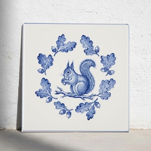 Op de afbeelding: Een vierkante keramische tegel met een blauw-witte illustratie van een eekhoorn op een eikentak. De eekhoorn is omringd door een cirkelvormige krans van gedetailleerde eikenbladeren en kleine eikels, geplaatst tegen een witte achtergrond.