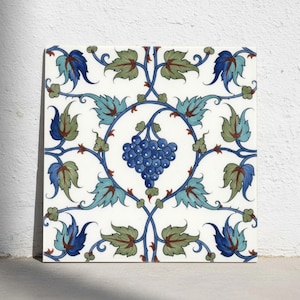 May include: A square ceramic tile featuring a central cluster of blue grapes framed by a circular vine pattern. The design includes symmetrical green and blue leaves with intricate swirling stems against a clean white background.