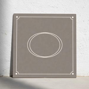 Puede incluir: Un letrero cuadrado de color topo con un borde blanco y un diseño de óvalo doble en el centro. Las esquinas del letrero tienen pequeños círculos blancos. El letrero está apoyado contra una pared blanca.