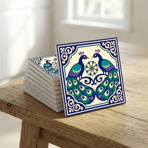 Könnte beinhalten: Ein Stapel quadratischer Keramikfliesen mit einem Pfauenmuster. Jede Fliese zeigt zwei sich zugewandte Pfauen mit türkisfarbenen, lila und grünen Federn und einem Blumenelement in der Mitte. Die Fliesen haben einen cremefarbenen Hintergrund und einen blauen Rand.