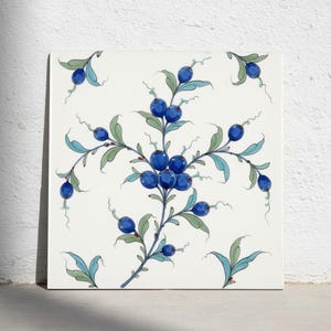 May include: A square ceramic tile featuring a hand-painted botanical pattern on a white background. The design displays a central cluster of dark blue berries on thin, branching stems with light green leaves and delicate, curling tendrils.