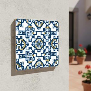 Puede incluir: Arte mural cuadrado con un patrón geométrico repetitivo en azul marino, dorado y blanco. El diseño incluye formas florales y geométricas estilizadas. La obra de arte está montada en una pared blanca, con una vista de un patio en el fondo.