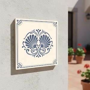 Puede incluir: Azulejo decorativo cuadrado, color crema, con un diseño de concha azul en el centro. El azulejo tiene un borde azul con detalles decorativos en las esquinas. Montado en una pared gris claro. Estilo mediterráneo clásico.
