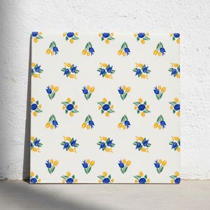 Puede incluir: Un azulejo de cerámica cuadrado con un patrón repetitivo de pequeños grupos de tulipanes pintados a mano. Cada grupo consta de dos tulipanes azules y uno amarillo con tallos y hojas verdes sobre un fondo de color blanco roto.