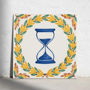 Könnte beinhalten: Eine quadratische Keramikfliese mit einer zentralen Abbildung einer blauen Sanduhr. Die Sanduhr ist von einem dekorativen Kranz aus gelben und grünen Blättern mit kleinen braunen Akzenten umgeben, alles auf einem cremeweißen Hintergrund.