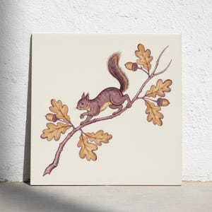 May include: A square decorative tile featuring a detailed illustration of a brown squirrel perched on an oak branch. The branch includes golden-yellow oak leaves and small brown acorns. The artwork is set against a solid off-white background.
