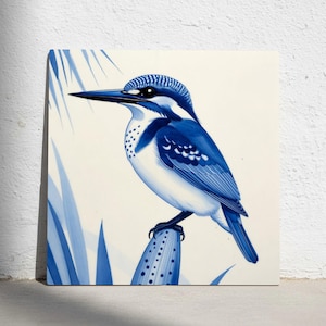 Op de afbeelding: Vierkant canvas kunstwerk met een blauw-witte illustratie van een ijsvogel die op een stengel zit. De vogel heeft gedetailleerde blauwe markeringen en staat tegen een witte achtergrond met blauwe bladeren. Het kunstwerk is aan een witte muur bevestigd.