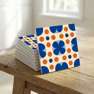 Puede incluir: Una pila de posavasos cuadrados de cerámica con un patrón repetitivo de formas azules y naranjas sobre un fondo crema. Los posavasos se muestran sobre una superficie de madera, con una ventana visible en el fondo.