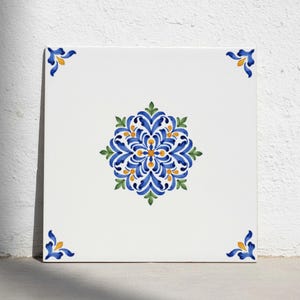 Op de afbeelding: Een vierkante witte keramische tegel met een centraal symmetrisch bloemmotief in blauw, geel en groen. Het ontwerp bestaat uit ingewikkelde wervelende bloemblaadjes en bladeren. Elk van de vier hoeken is geaccentueerd met een klein blauw en geel bloemelement.