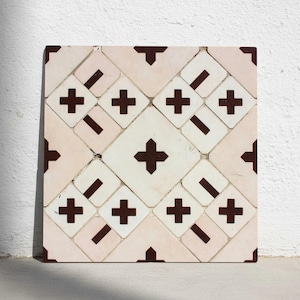Op de afbeelding: Vierkante tegel met een herhalend patroon van witte vierkanten en lichtroze rechthoeken. De witte vierkanten hebben een donkerbruin kruisontwerp, terwijl de rechthoeken een donkerbruine diagonale lijn hebben. De hoeken van de tegel zijn ook donkerbruin.